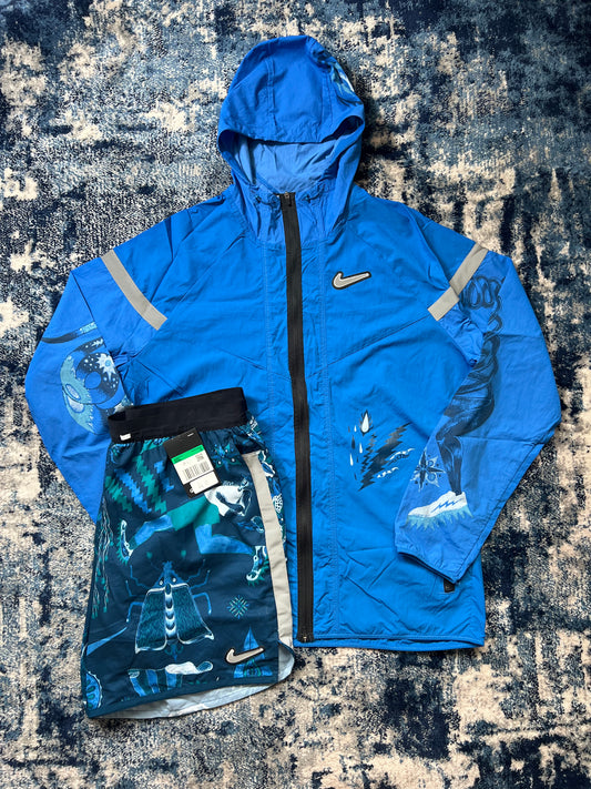 A blue Nike rain jacket with reflective stripes and a black zipper, laid flat next to a pair of blue and black patterned Nike. Blue Dragon set shorts on a dark textured rug.