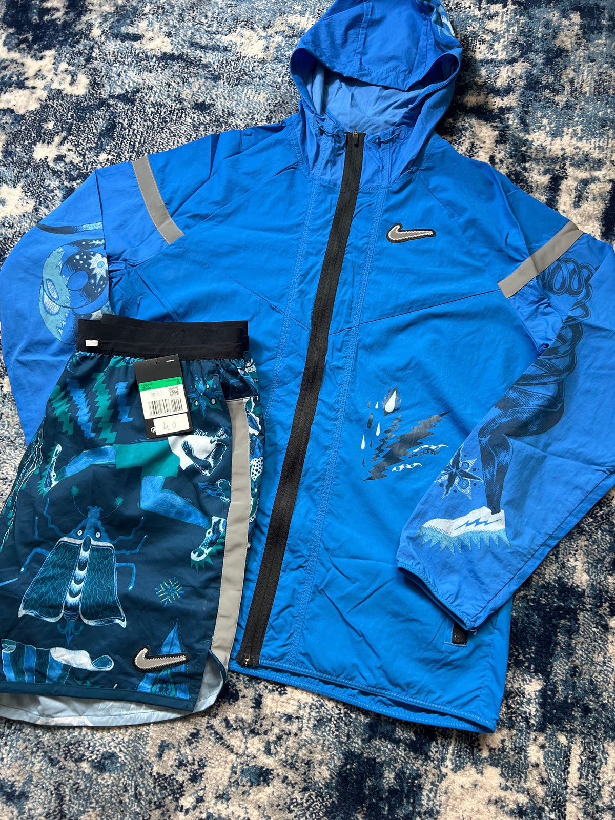A blue Nike hooded jacket and matching printed shorts laid flat on a textured gray and white rug. The Nike logo is visible on both items, and the shorts feature a dark blue, green, and white graphic print.